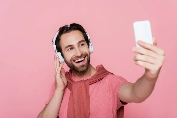 Hombre sonriente en auriculares tomando selfie en un teléfono inteligente borroso aislado en rosa - foto de stock