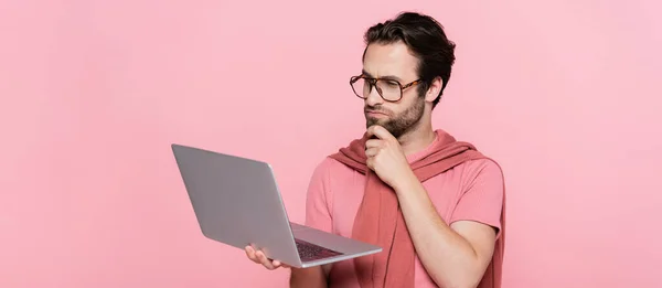 Thoughtful man in eyeglasses looking at laptop isolated on pink, banner — Stock Photo