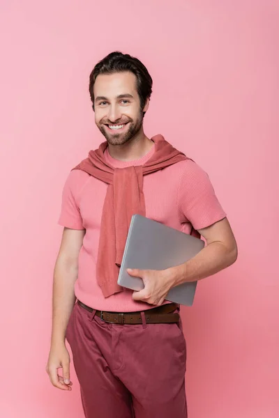 Смолящий фрилансер с джинсами на плечах, держащий ноутбук изолированным на розовом — стоковое фото