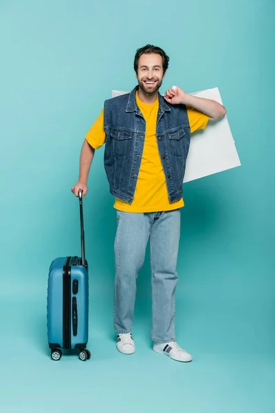 Hombre positivo sosteniendo bolsa de compras y maleta sobre fondo azul - foto de stock