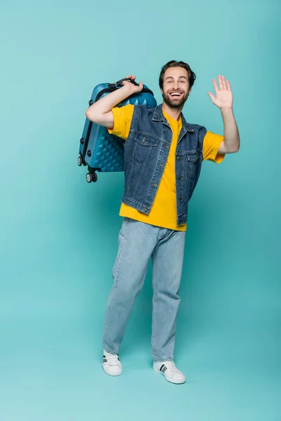 Viajante feliz em colete jeans segurando mala e acenando mão no fundo azul — Fotografia de Stock