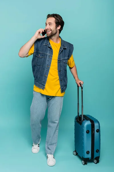 Turista sonriente hablando por teléfono móvil y sosteniendo la maleta sobre fondo azul - foto de stock