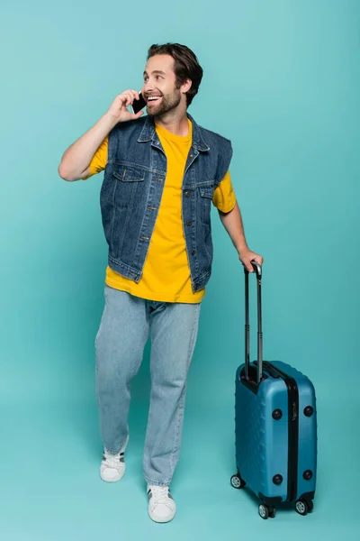 Turista sonriente hablando por teléfono móvil y sosteniendo la maleta sobre fondo azul — Stock Photo