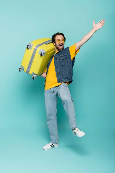 Turista excitado en gafas de sol sosteniendo maleta sobre fondo azul - foto de stock