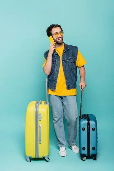 Homme souriant en lunettes de soleil parlant sur smartphone près de valises sur fond bleu — Photo de stock