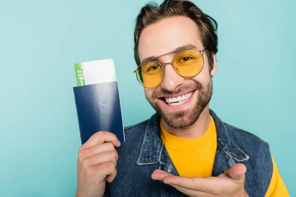 Smiling man in sunglasses pointing at passport and air ticket isolated on blue — Stock Photo