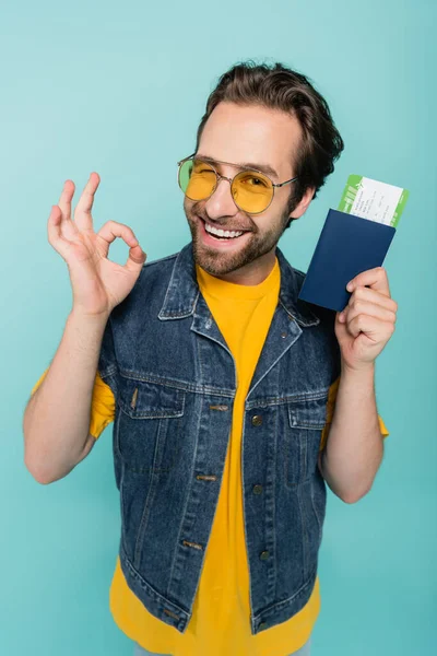 Lächelnder Mann zeigt OK-Geste, während er Pass und Flugticket isoliert auf blauem Grund hält — Stockfoto