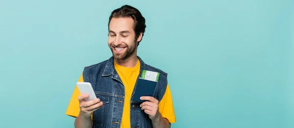 Homem positivo segurando smartphone e passaporte isolado em azul, banner — Fotografia de Stock