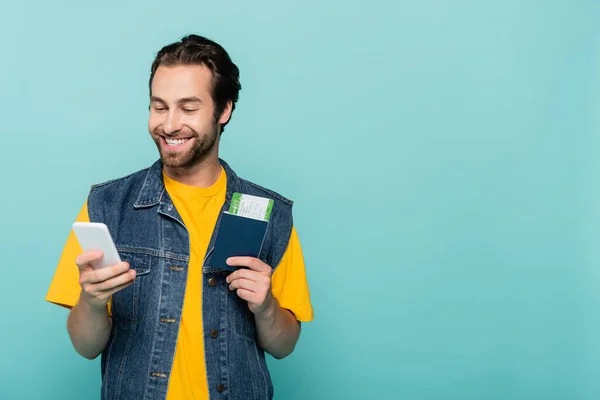 Homem sorridente segurando smartphone e passaporte com cartão de embarque isolado em azul — Fotografia de Stock