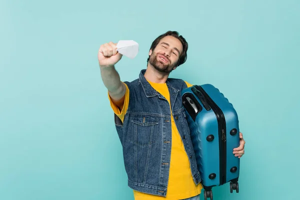 Homem positivo segurando mala e brincando com o plano de papel isolado em azul — Fotografia de Stock