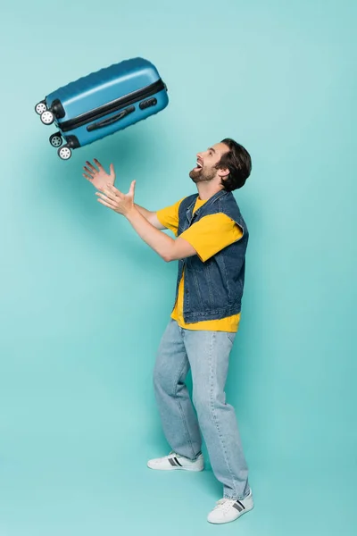 Side view of cheerful man throwing suitcase on blue background — Stock Photo