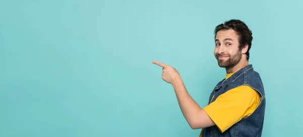 Uomo sorridente in t-shirt e gilet in denim puntato con dito isolato su blu, striscione — Foto stock