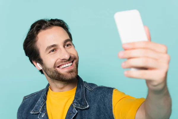 Homme souriant dans l'écouteur prendre selfie sur smartphone isolé sur bleu — Photo de stock