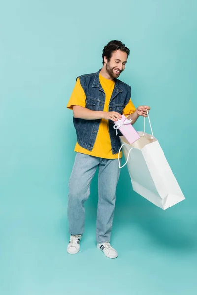 Junger Mann steckt Geschenk in Einkaufstasche auf blauem Hintergrund — Stockfoto