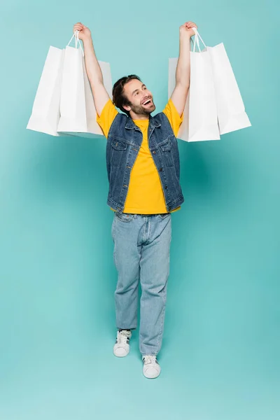 Full length of smiling man rising shopping bags on blue background — Stock Photo