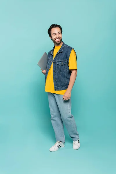 Longitud completa de teletrabajador sonriente sosteniendo el ordenador portátil sobre fondo azul - foto de stock