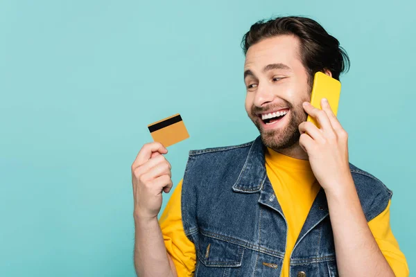 Sonriente hombre hablando en el teléfono móvil y mirando a la tarjeta de crédito aislado en azul - foto de stock