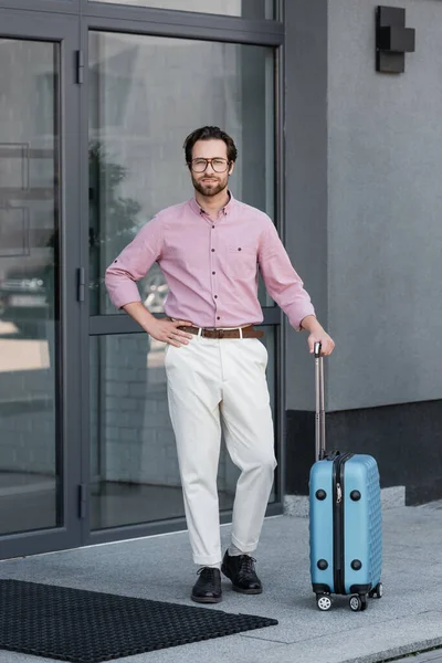 Uomo d'affari sorridente con valigia in piedi vicino all'edificio — Stock Photo