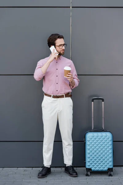 Joven hombre de negocios con taza de papel hablando en teléfono inteligente cerca de la pared - foto de stock