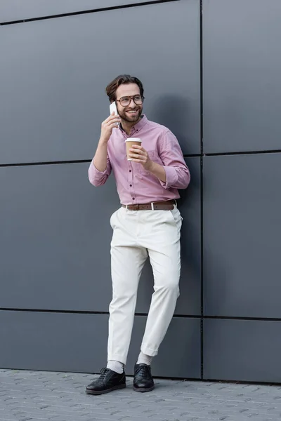 Empresário alegre falando no telefone celular e segurando copo de papel perto do edifício — Fotografia de Stock