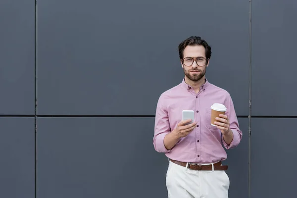 Joven hombre de negocios con taza de papel y teléfono inteligente mirando a la cámara cerca del edificio — Stock Photo