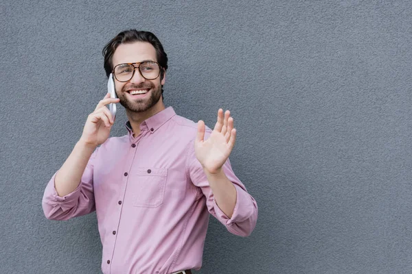 Hombre de negocios sonriente hablando en smartphone y saludando con la mano - foto de stock