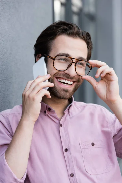 Hombre de negocios sonriente sosteniendo gafas mientras habla en el teléfono inteligente cerca del edificio — Stock Photo