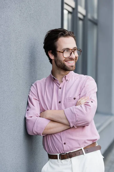 Smiling businessman in eyeglasses standing with crossed arms near building — Stock Photo