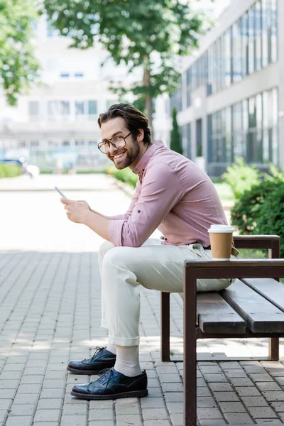 Uomo d'affari sorridente utilizzando smartphone vicino al caffè per andare in panchina — Foto stock