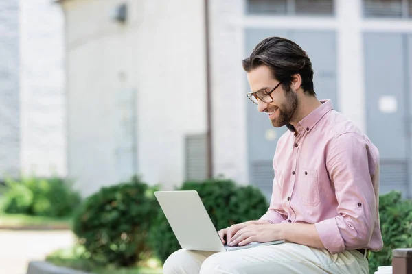 Side view of businessman in eyeglasses smiling while using laptop outdoors — Stock Photo