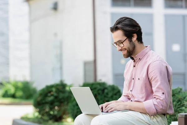 Vista laterale dell'uomo d'affari con gli occhiali che sorride mentre usa il computer portatile all'aperto — Foto stock