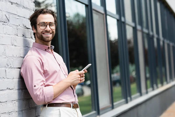 Giovane manager che sorride alla fotocamera e tiene lo smartphone vicino all'edificio — Foto stock