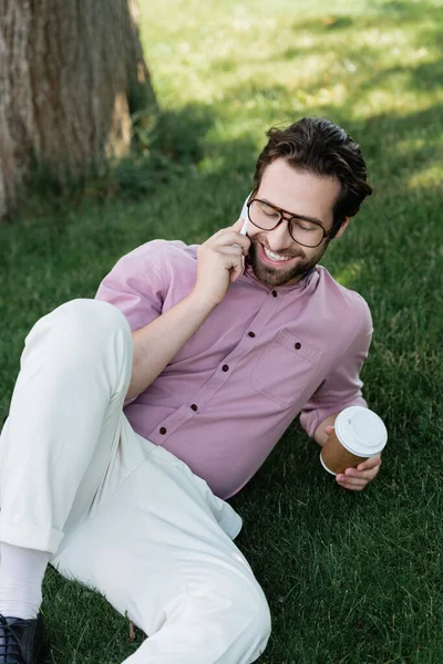 Cheerful businessman talking on smartphone and holding paper cup on grass — Stock Photo