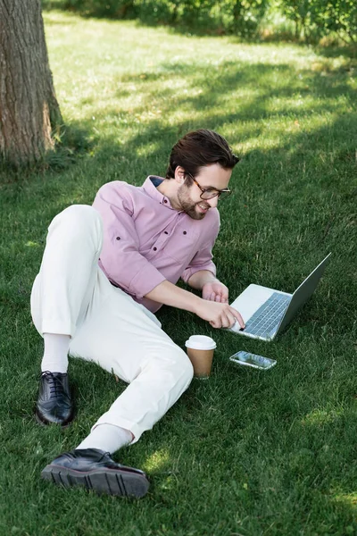 Vista de ángulo alto del hombre de negocios sonriente usando el ordenador portátil cerca de la bebida para llevar y el teléfono inteligente en la hierba - foto de stock
