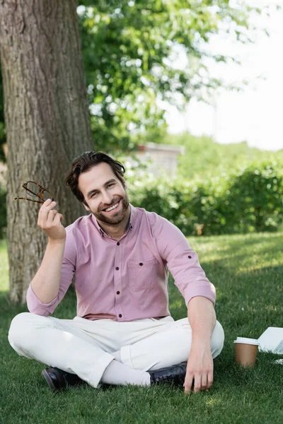 Geschäftsmann hält Brille neben Laptop und Pappbecher auf Gras — Stockfoto