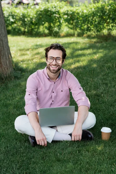 Hombre de negocios positivo con portátil mirando a la cámara cerca de la taza de papel en la hierba - foto de stock