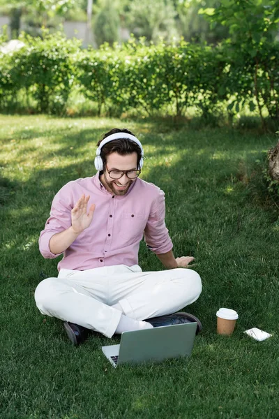 Hombre de negocios sonriente en auriculares que tienen videollamada en el portátil cerca de la taza de papel en la hierba en el parque - foto de stock