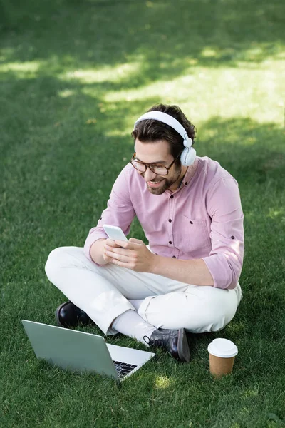 Young businessman in headphones using devices near coffee to go in park — Stock Photo