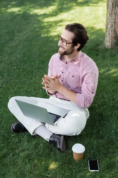 Homme d'affaires souriant avec ordinateur portable regardant loin près de téléphone portable et café dans le parc — Photo de stock