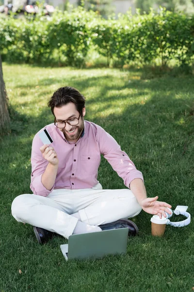 Hombre de negocios sonriente con tarjeta de crédito que tiene videollamada en el portátil cerca de los auriculares y bebida para llevar en el parque - foto de stock
