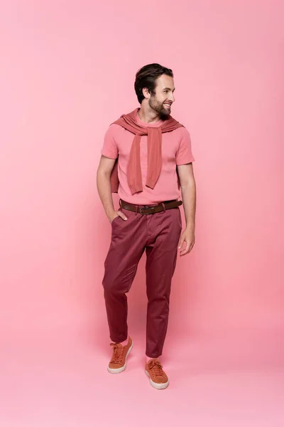 Longitud completa del hombre sonriente sosteniendo la mano en el bolsillo de los pantalones sobre fondo rosa - foto de stock