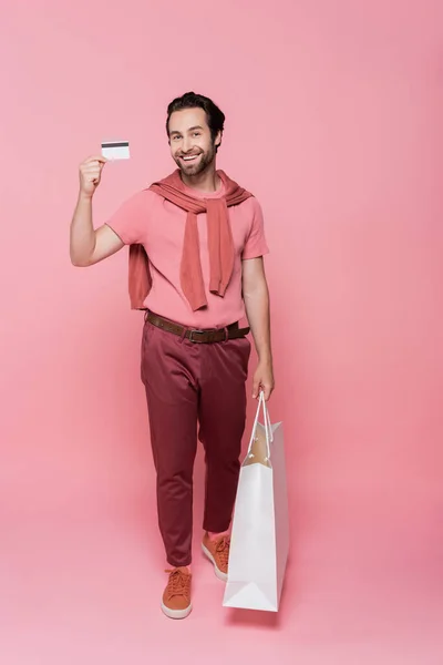 Positive man holding credit card and white shopping bag on pink background — Stock Photo