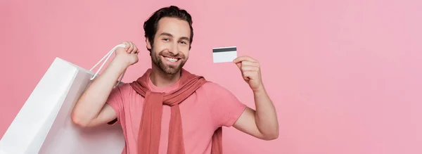 Чоловік з сумкою, що показує кредитну картку ізольовано на рожевому, банер — стокове фото