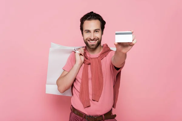 Розмита кредитна картка в руці усміхненого чоловіка з сумкою для покупок ізольована на рожевому — стокове фото
