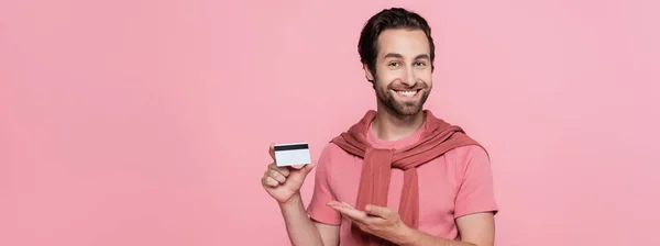 Uomo positivo che punta con mano la carta di credito isolata su rosa, banner — Foto stock