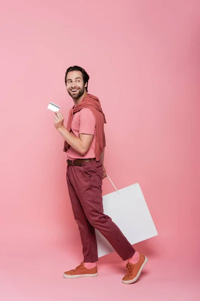 Shopaholic in voller Länge mit Kreditkarte und Einkaufstasche auf rosa Hintergrund — Stockfoto