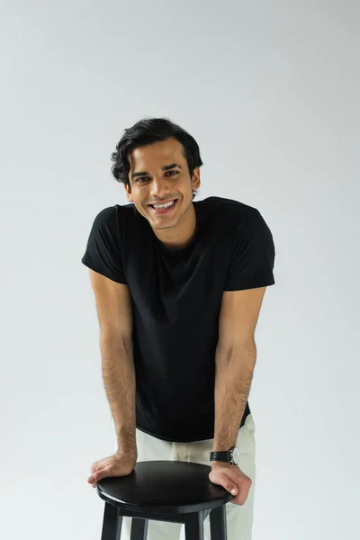 Portrait of cheerful young man looking at camera and leaning on chair isolated on grey — Stock Photo