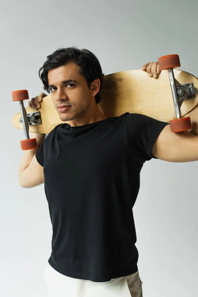 Jeune homme en t-shirt noir tenant longboard et regardant la caméra isolée sur gris — Photo de stock