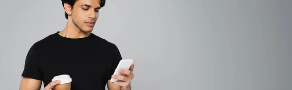 Young man holding coffee to go and using smartphone isolated on grey, banner — Stock Photo