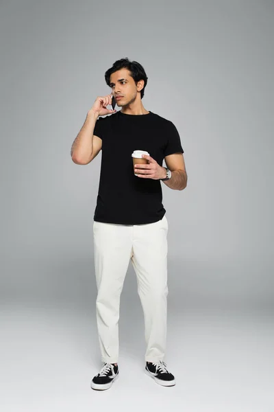 Full length of young man holding coffee to go and talking on smartphone on grey — Stock Photo